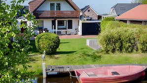 A residential area with a house, a boat docked at a pier, and a well-maintained lawn. - Holiday Home for 5 People (105 m²) in Südbrookmerland (Südbrookmerland)