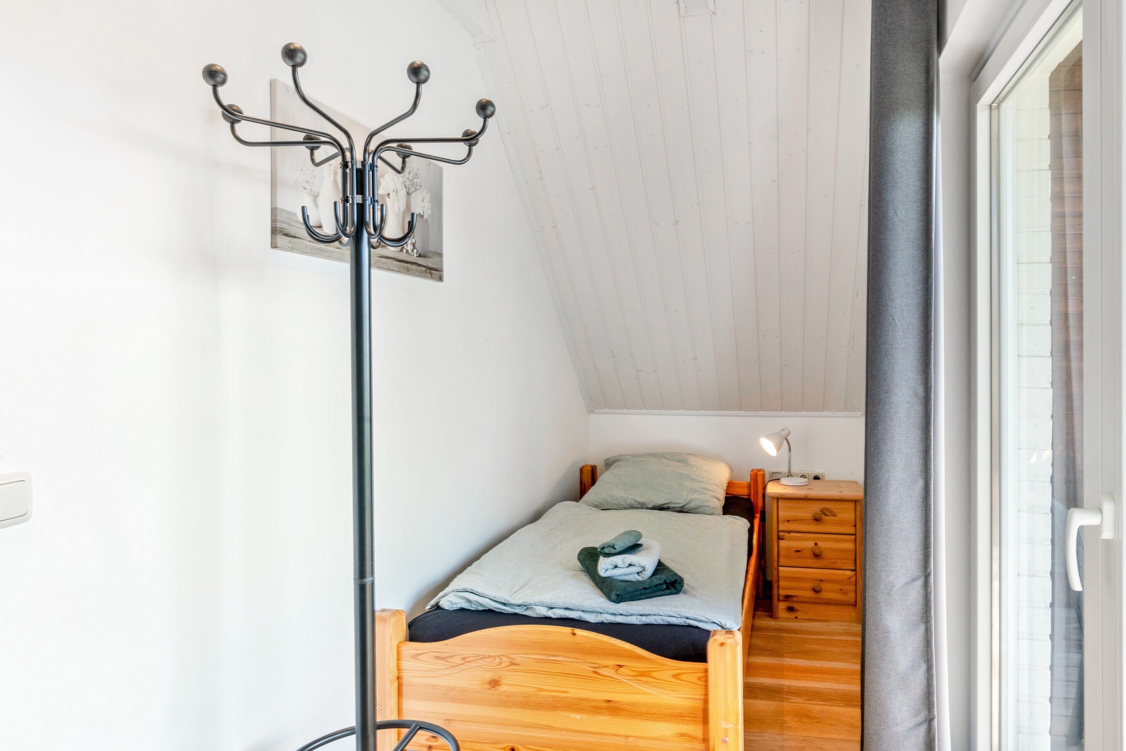 A small, cozy room with a wooden bed, a nightstand, a lamp, and a coat rack.