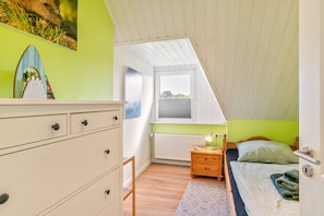 A bedroom with a sloped ceiling, a bed with a green pillow, a nightstand with a lamp, a dresser, and a window with a view. - Holiday Home for 5 People (105 m²) in Südbrookmerland (Südbrookmerland)