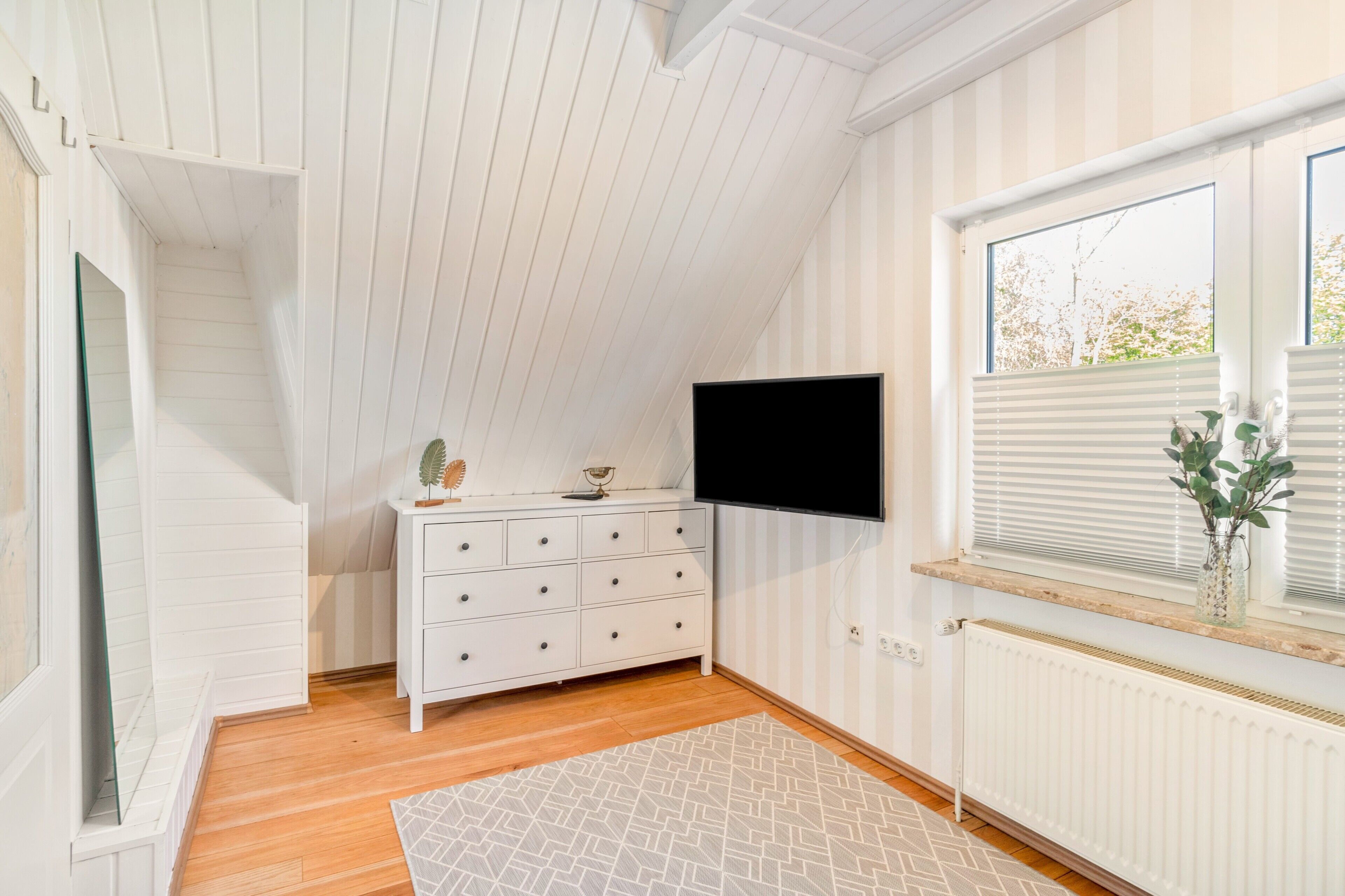 Una habitación con techo inclinado, un aparador blanco, un televisor de pantalla plana montado en la pared, una ventana con persianas y un radiador.