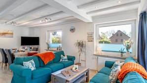 A living room with teal sofas, a coffee table, and a TV. There is a dining area with chairs and a table. - Holiday Home for 5 People (105 m²) in Südbrookmerland (Südbrookmerland)