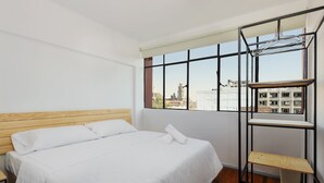 Ein Schlafzimmer mit einem Bett, einem Fenster mit Blick auf Gebäude und einem Metallregal.