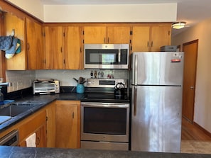 A kitchen with wooden cabinets, a microwave, a toaster, a kettle, and a refrigerator. - Iris Cottage-Walk to Short Sands (York)