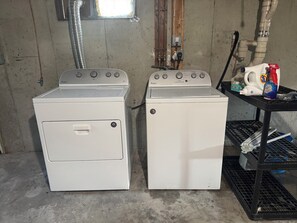 Two white Whirlpool appliances, a gas stove and a dryer, in a room with concrete walls and a window. - Iris Cottage-Walk to Short Sands (York)