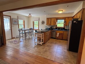 A kitchen with wooden cabinets, a refrigerator, and a dining area with chairs and a table. - Iris Cottage-Walk to Short Sands (York)