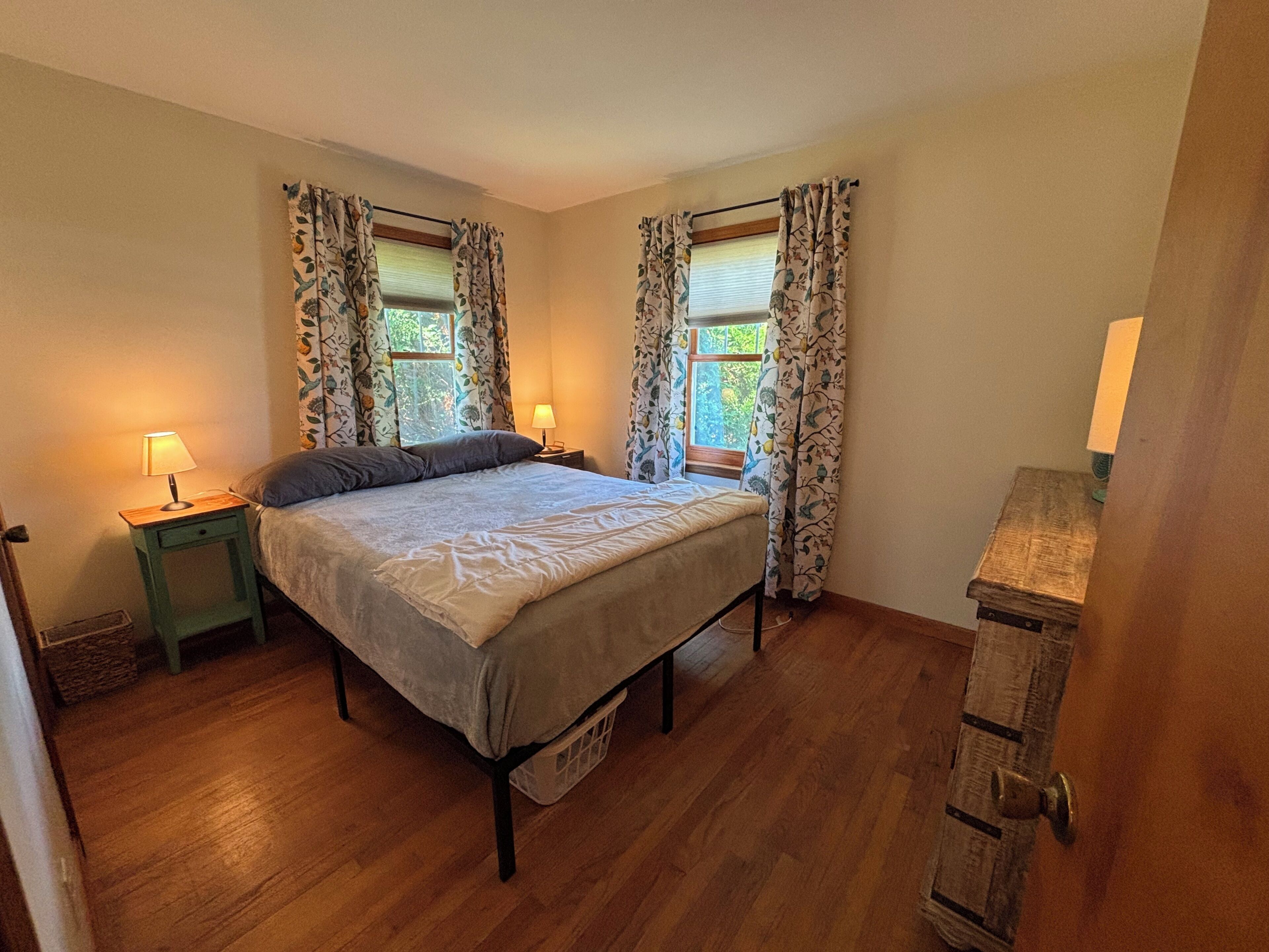 Een slaapkamer met een bed, twee bedlampen, een houten nachtkastje en een raam met gordijnen.