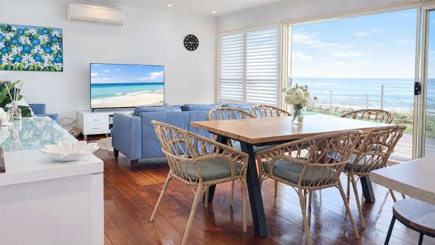 Un salon moderne comprenant un canapé, un téléviseur, une table à manger, des chaises en osier et une vue sur l’océan.