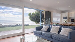 Un salon moderne avec une table basse en verre, un canapé bleu agrémenté de coussins et une vue sur l’océan grâce à de grandes portes coulissantes.
