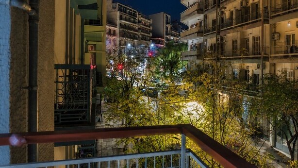 Vista nocturna de un edificio residencial de varios pisos con balcones, una entrada protegida por un portón y una calle con farolas y automóviles estacionados.