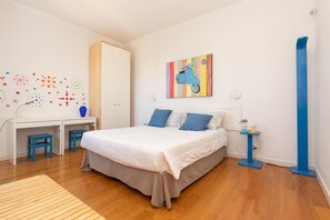 Ein Schlafzimmer mit einem Bett, einem Schreibtisch, zwei Stühlen, einem Kleiderschrank, einer farbenfrohen Wanddekoration und einer blauen Stehlampe.