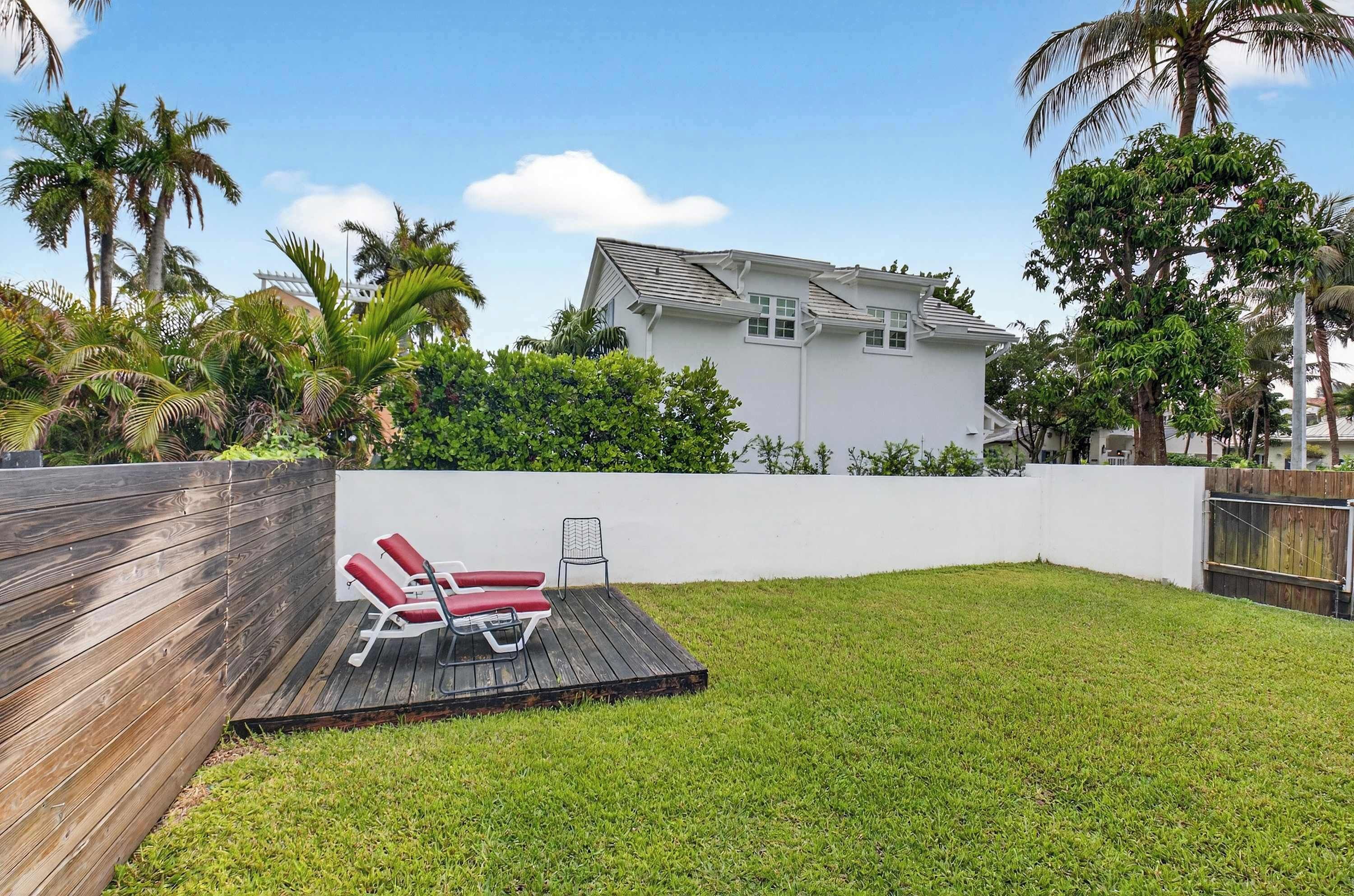 A backyard with a wooden deck, two red lounge chairs, a white wall, and a white house in the background.