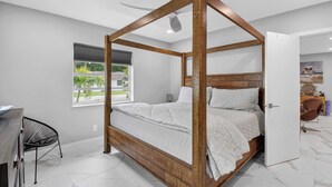 Un lit à baldaquin avec un cadre en bois, un ventilateur de plafond, une fenêtre avec des stores et une vue sur la verdure à l’extérieur.