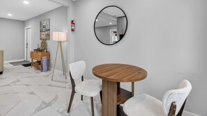 Une chambre d’hôtel avec une table ronde en bois, deux chaises blanches à revêtement, un miroir rond, une lampe sur pied et un petit meuble contenant des objets décoratifs.