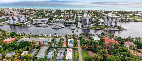 An aerial view of a coastal marina with multiple boats docked, surrounded by residential buildings and lush greenery.
