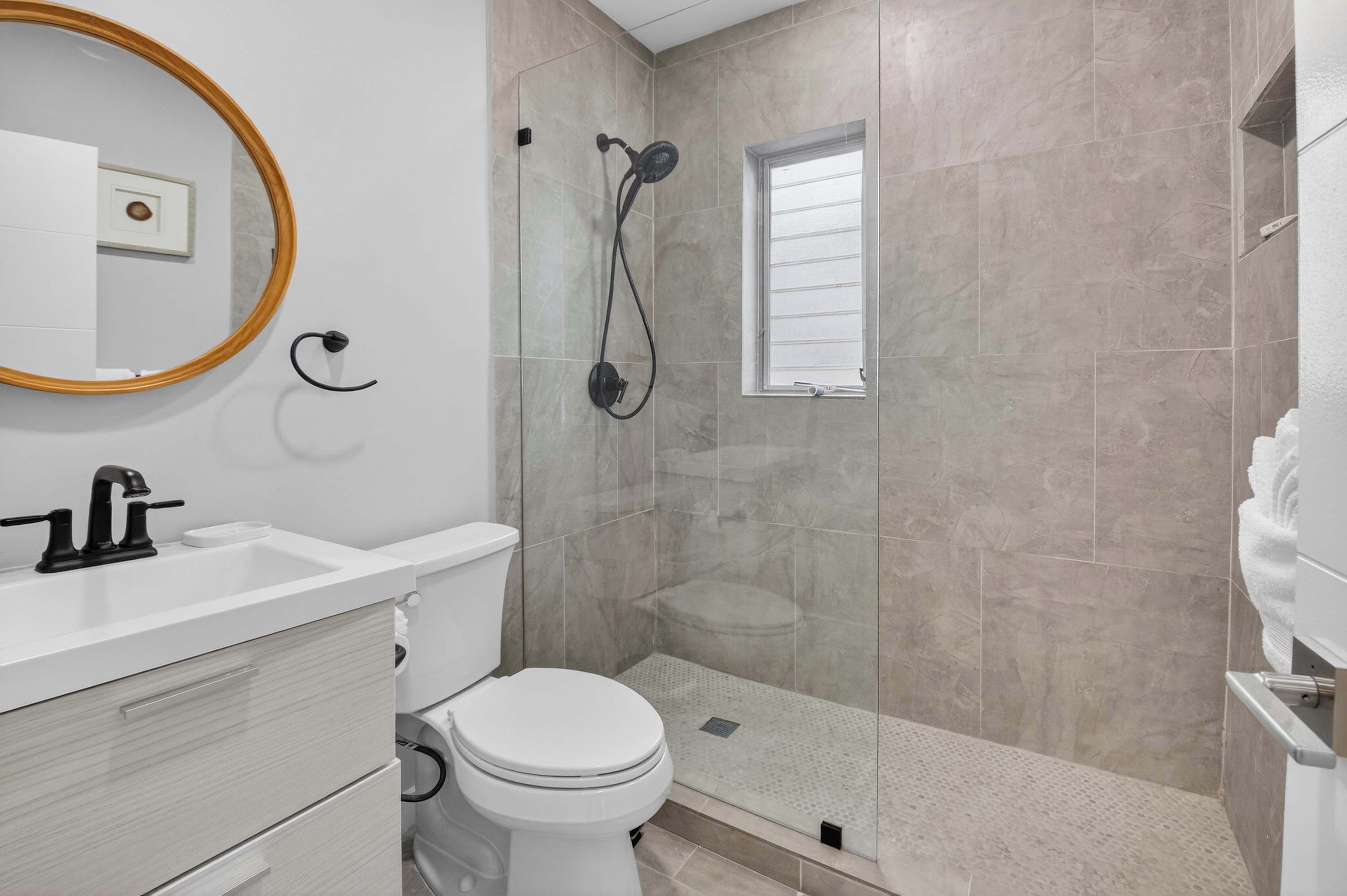 A bathroom with a toilet, sink, mirror, shower, and a window.