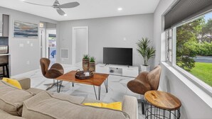 Un salon moderne doté d’un ventilateur de plafond, d’un téléviseur à écran plat, d’une table à manger avec des chaises et d’une grande fenêtre donnant sur un paysage verdoyant.