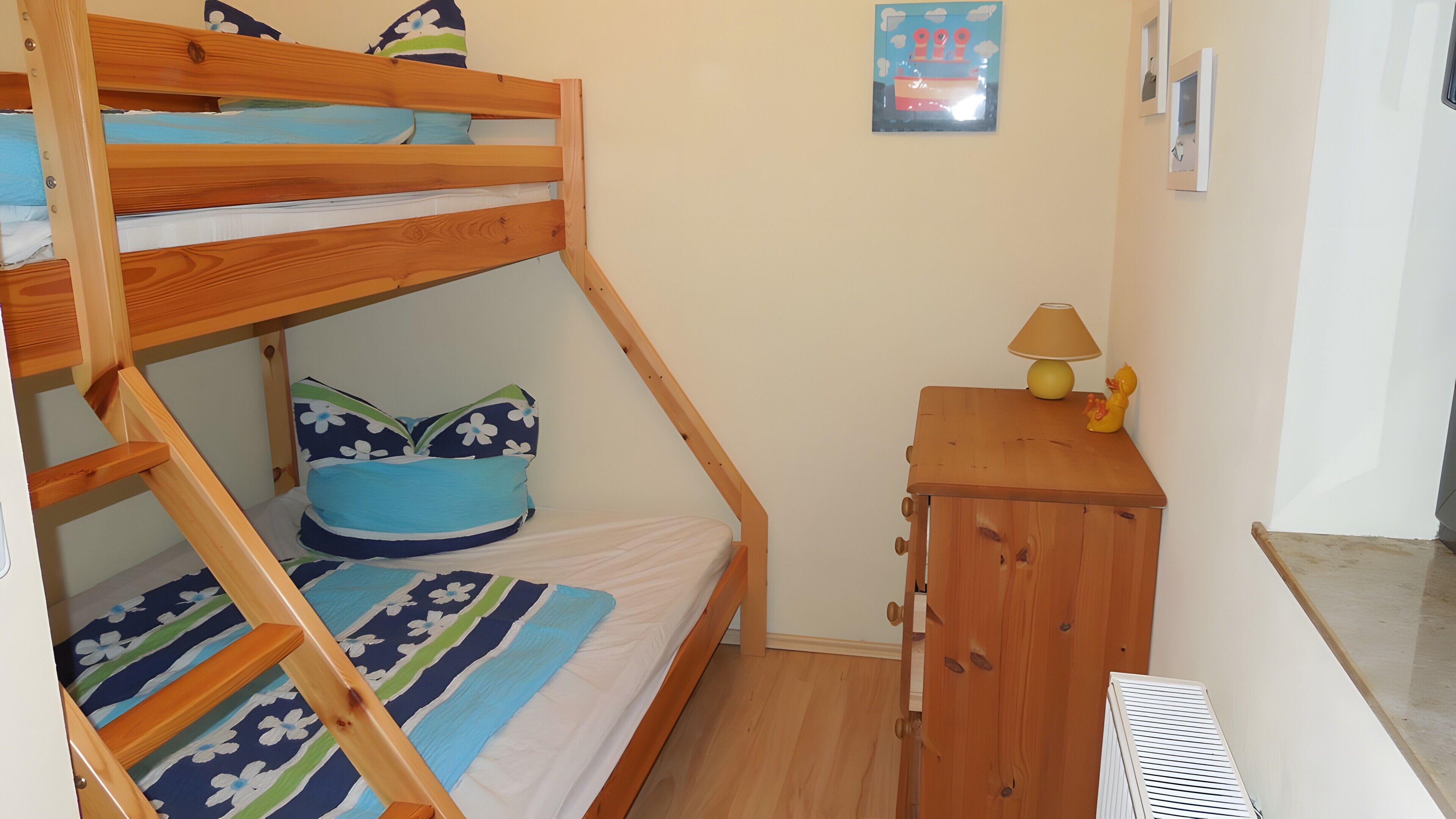A bunk bed with a blue floral bedspread, a small wooden nightstand with a lamp, and a wall-mounted picture.