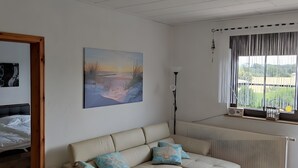 Une pièce avec un canapé beige, un tableau représentant un coucher de soleil, un radiateur et une fenêtre avec des rideaux.