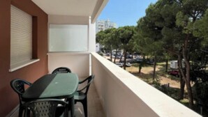 Een balkon met een tafel en stoelen, een raam met zonwering en uitzicht op bomen en gebouwen.