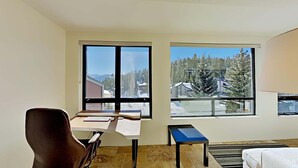 A room with a desk, chair, and a view of a snowy landscape through large windows. - 1849 Condos 309 2 Bedroom Condo (Mammoth Lakes)