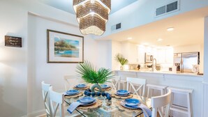 A dining area with a glass table, white chairs, a chandelier, a framed painting, and a kitchen in the background. - Oceanfront Penthouse w/ Outdoor Balconies, Community Pool, Beach Access, Playground & Kid's Pool (Isle of Palms)