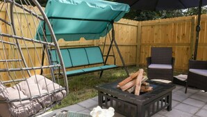 Une terrasse avec un fauteuil suspendu, une table avec un saladier et un foyer.