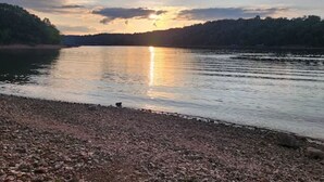 Un lac paisible au coucher du soleil, avec une rive de galets et des collines boisées en arrière-plan.