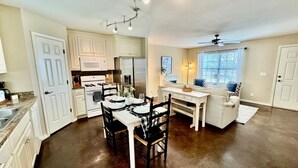 Une cuisine avec des meubles blancs, une table à manger avec des chaises, un ventilateur de plafond et une fenêtre avec des stores.