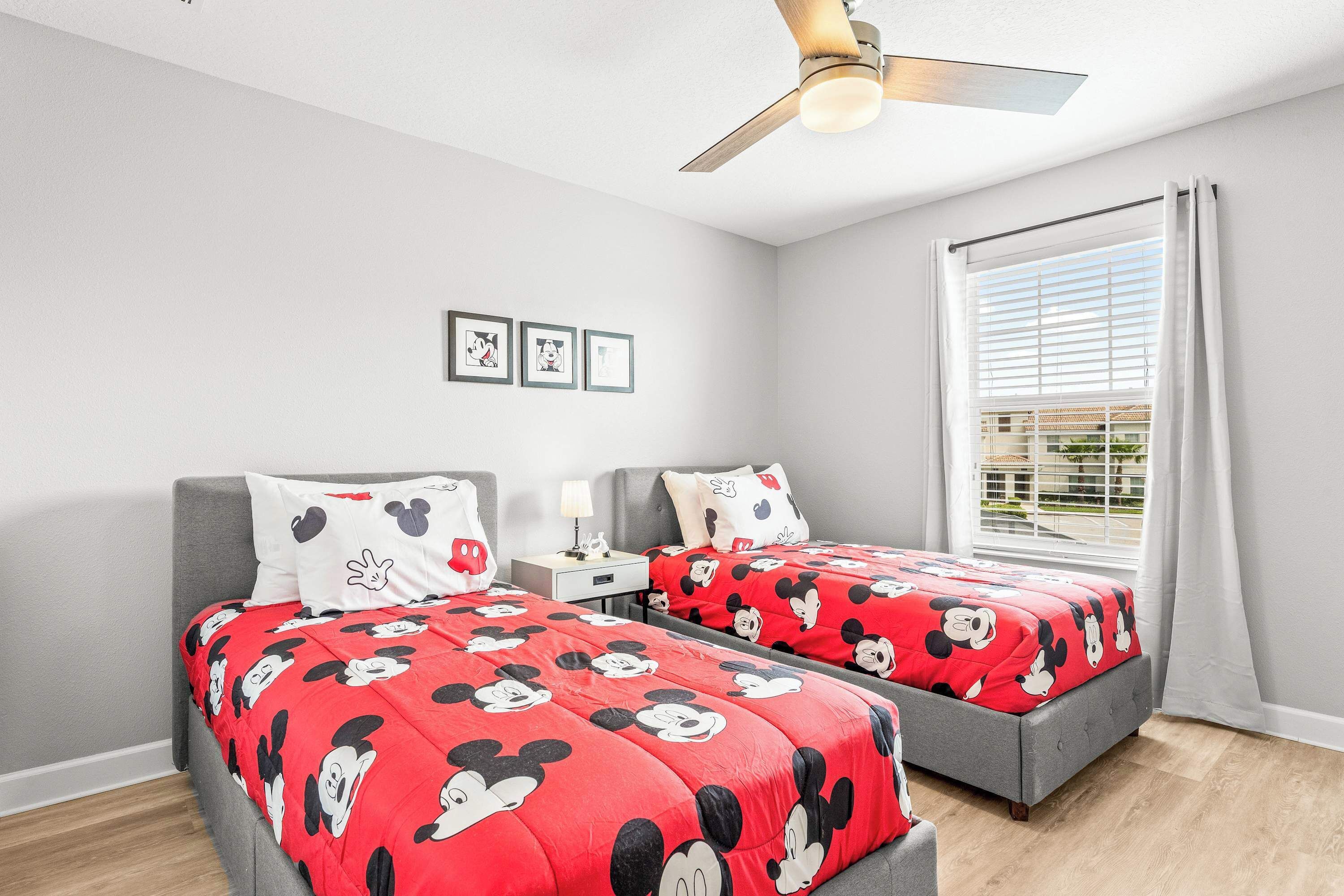 A bedroom with two beds, a ceiling fan, a window with curtains, and framed pictures on the wall.