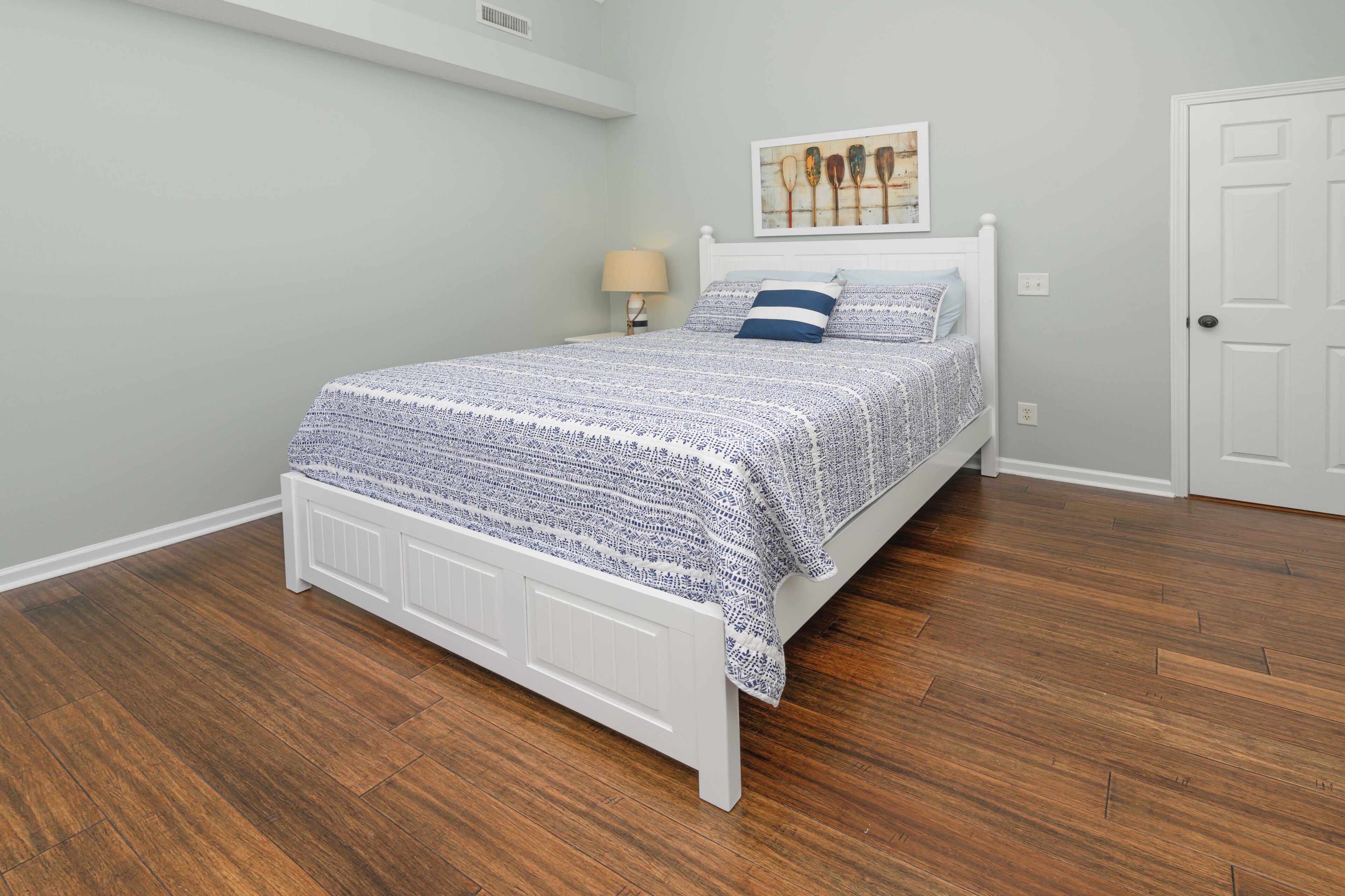Quarto com cama branca, mesa de cabeceira com abajur, quadro na parede e uma porta.