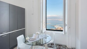 A modern room with a glass table, white chairs, a view of the sea, and grey cabinets. - Apartment 'La Finestra' with Sea View, Wi-Fi and Air Conditioning (Castellammare di Stabia)