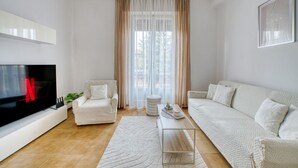Ein modernes Wohnzimmer mit Flachbildfernseher, zwei weißen Sofas, einem Couchtisch und einem großen Fenster mit transparenten Vorhängen.