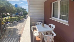 Un balcón con una mesa y sillas, una ventana y vista a una calle con coches aparcados y árboles.
