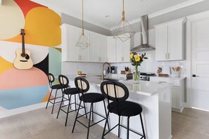 Eine moderne Küche mit weißen Schränken, einer zentralen Kochinsel mit Barhocker, Pendelleuchten und einer Gitarre an der Wand.
