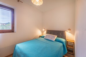 A bedroom with a bed, a nightstand, a window, and a wall-mounted light fixture. - Villa Filippa Pittulongu – Cozy Holiday Home Near the Sea for 4 Guests (Pittulongu)