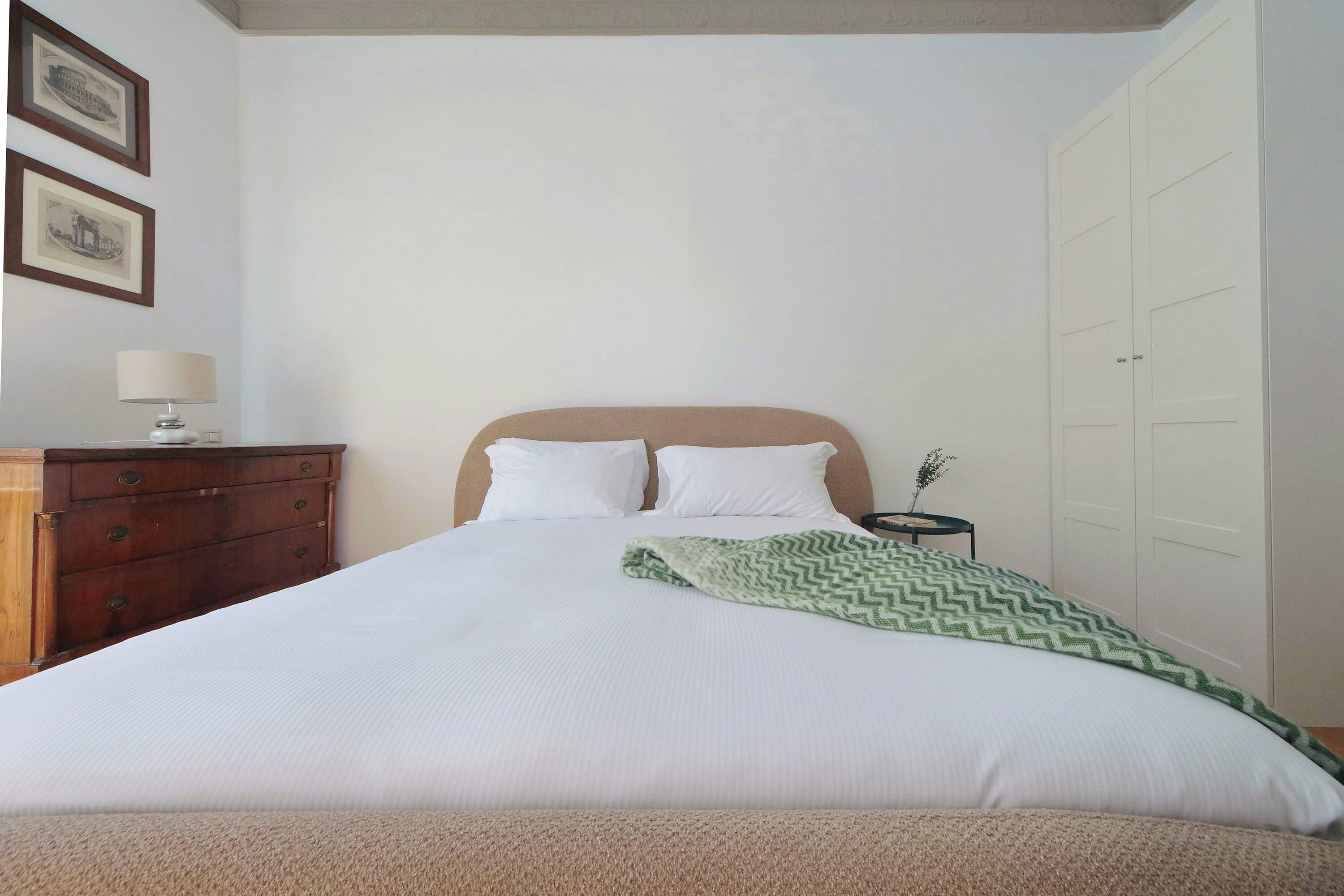 Ein ordentlich gemachtes Bett mit einer grün gemusterten Decke, eine hölzerne Kommode mit einer Lampe und zwei gerahmte Bilder an der Wand.