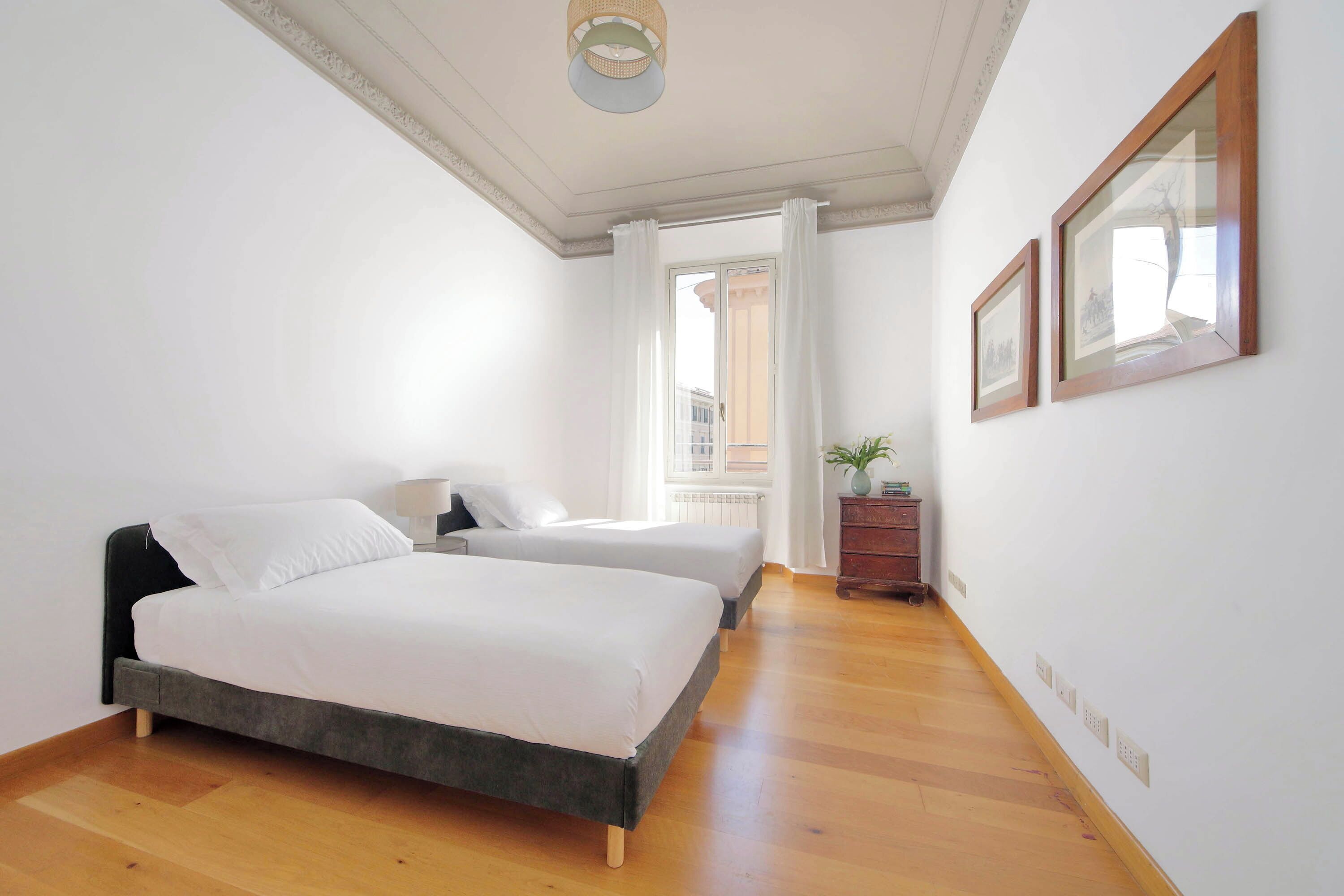 A bedroom with two beds, a wooden floor, a window with curtains, and a small plant on a dresser.