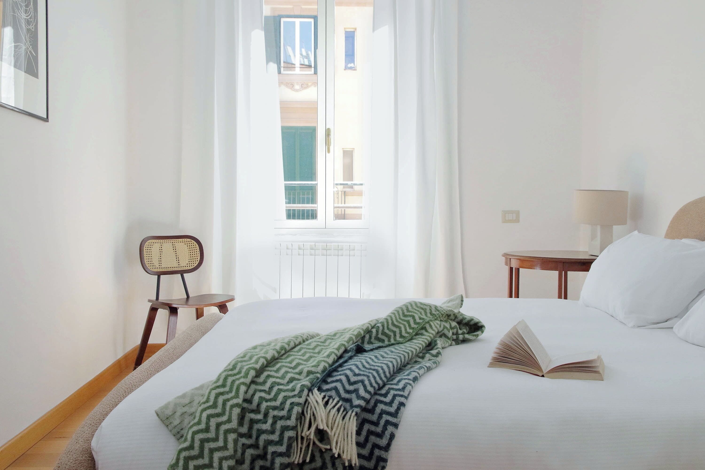 A hotel room with a bed, a chair, a table, and a view of the outside.