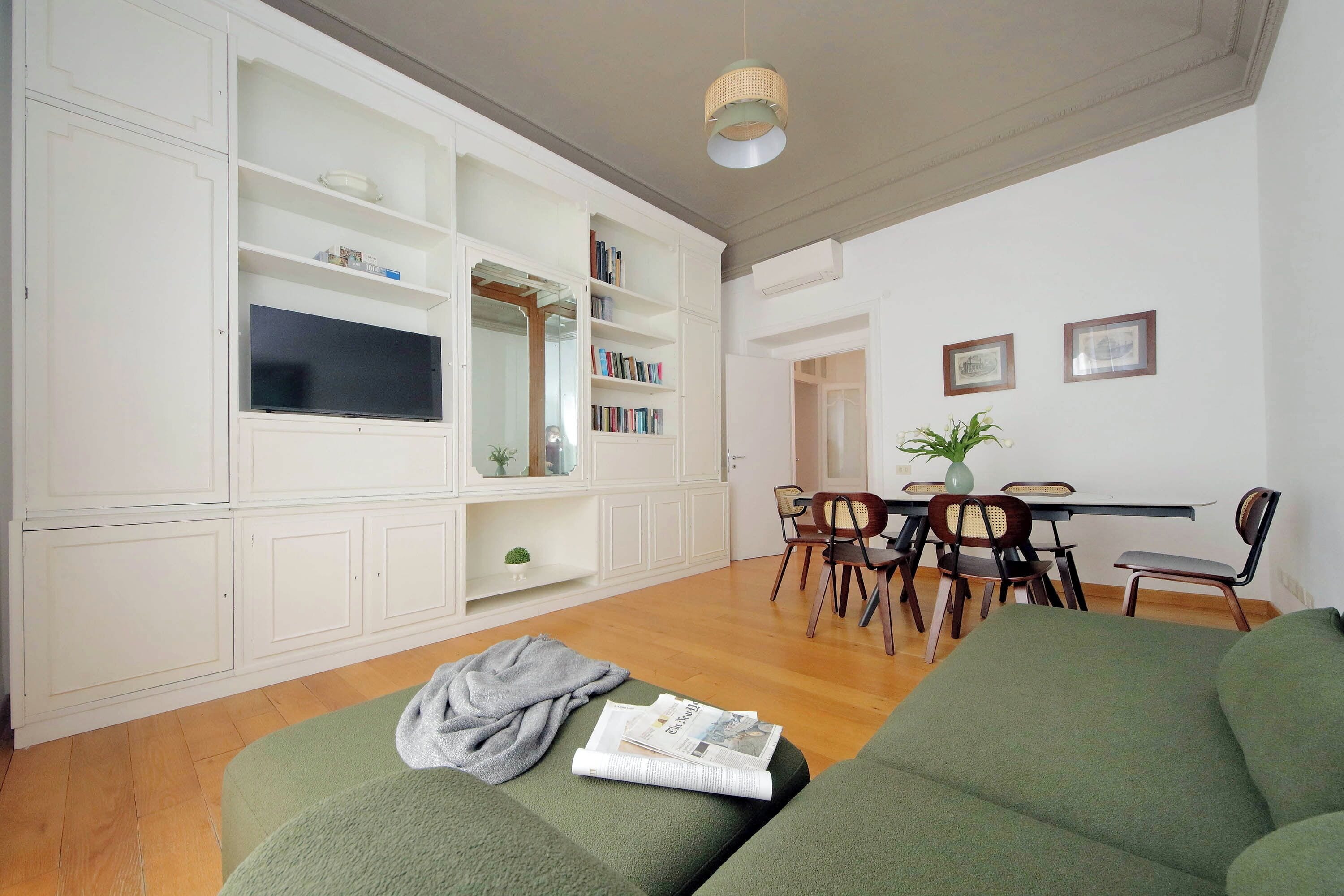Ein Wohnzimmer mit grünem Sofa, einem Flachbildfernseher, weißen Einbauschränken, einem Essbereich mit Tisch und Stühlen sowie einer Pendelleuchte.