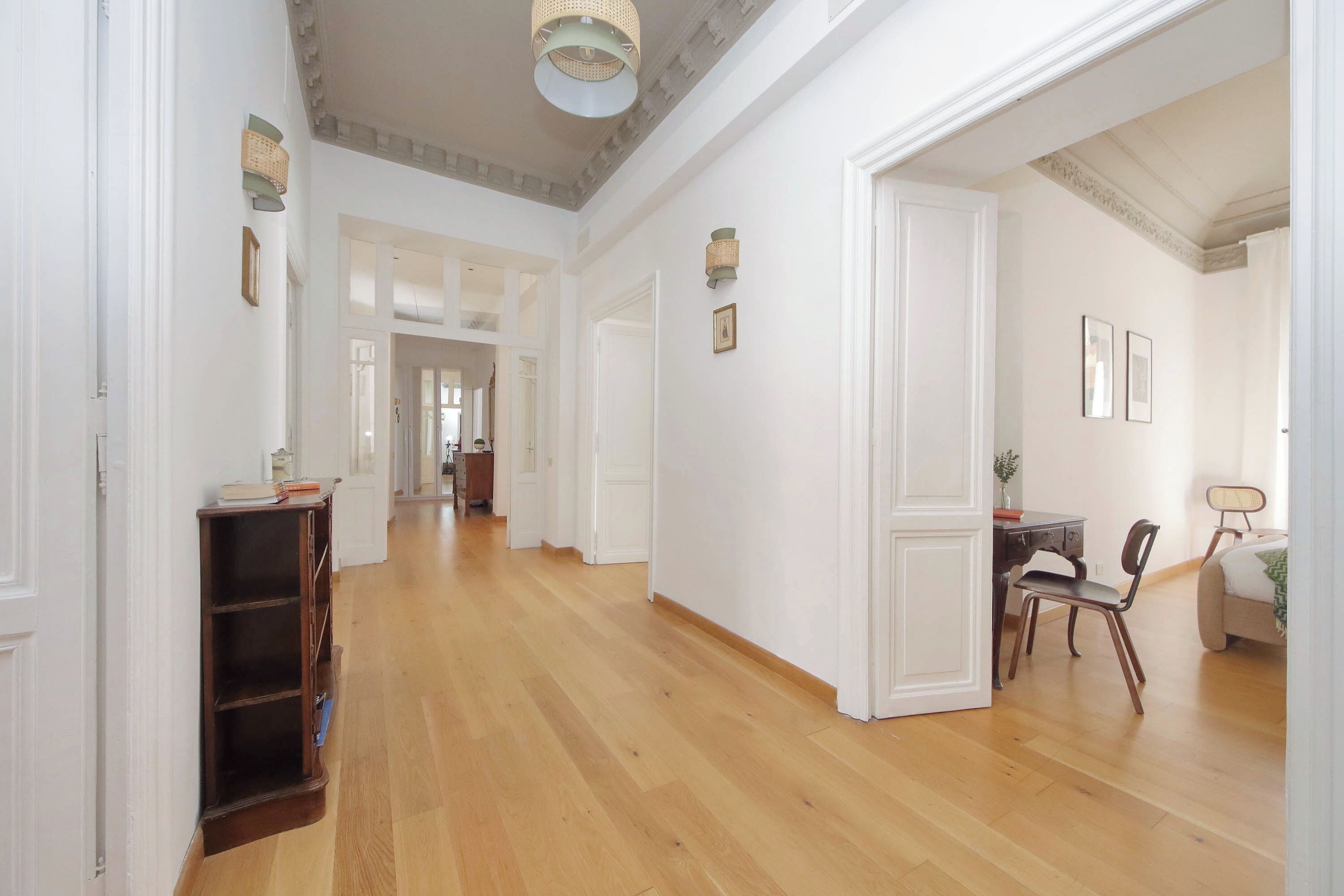 A spacious hallway with wooden flooring, white walls, and a ceiling with exposed beams. There are doors leading to other rooms, a small table with chairs, and wall-mounted lamps.