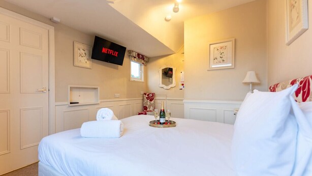 A hotel room with a bed, a television mounted on the wall, a mirror, and a bedside table with a bottle and glasses.