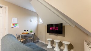 A modern living room with a grey sofa, a dining table with two chairs, a TV mounted on the wall, and three small white stools. - Cransley Room 11 - Apartment close to the beach with communal garden (Bournemouth)