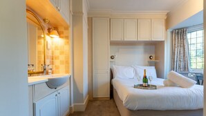 Une chambre à coucher avec un lit, une table de chevet sur laquelle se trouvent une bouteille et des verres, une fenêtre avec des rideaux, et une salle de bain avec un lavabo et un miroir.