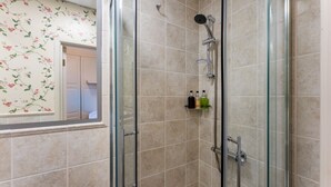 Une douche fermée par des parois de verre, avec des carreaux de couleur beige, un pommeau de douche et une étagère contenant des articles de toilette.