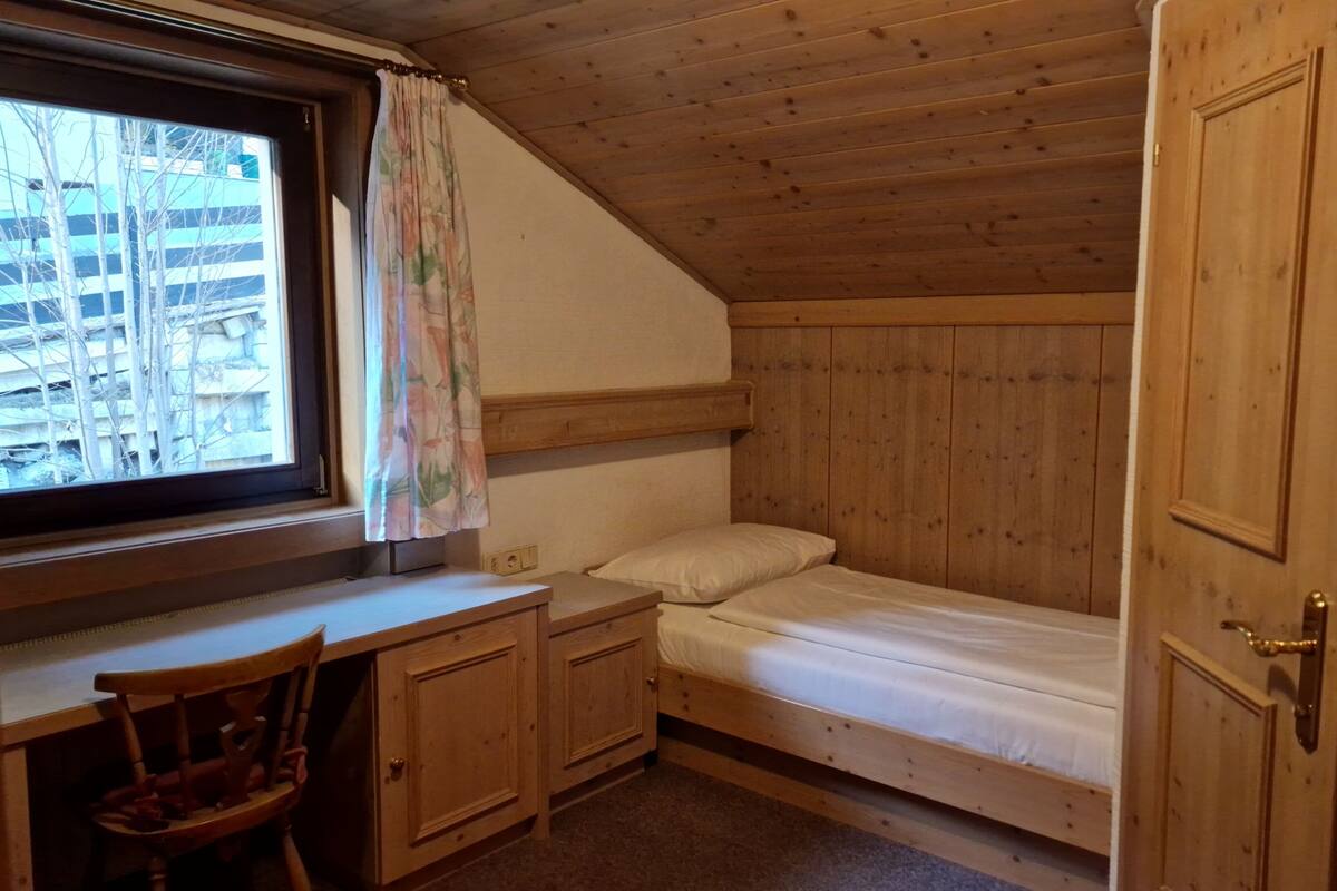 Eine kleine, einräumige Holzhütte mit Holzbalkendecke, einem Schreibtisch mit Stuhl, einem Bett und einem Fenster mit Vorhang.