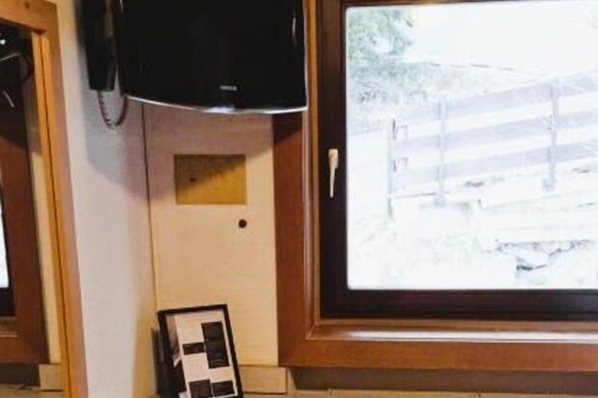 Ein Zimmer mit einem Holztisch, einem Sessel, einem an der Wand befestigten Fernseher und einem Fenster mit Blick auf einen Zaun.