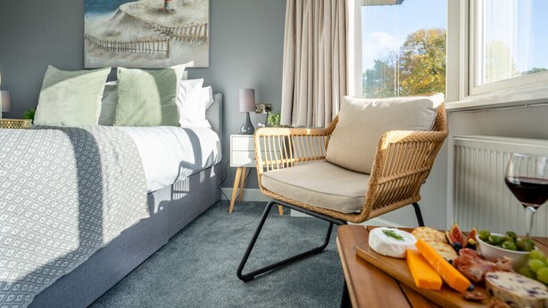A bedroom with a bed, a wicker chair, a nightstand, a lamp, a window with curtains, and a table with food. - Cransley Room 6 - Close to beaches and town with shared garden (Bournemouth)