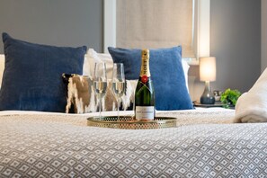 Ein Bett mit einer Champagnerflasche und Gläsern, einer gemusterten Tagesdecke und blauen Kissen.