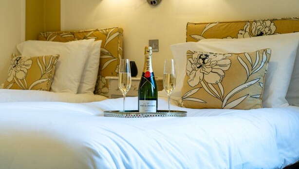 Ein Bett mit floralen Kissen, ein Tablett mit einer Champagnerflasche und zwei Gläsern sowie eine Wandleuchte.
