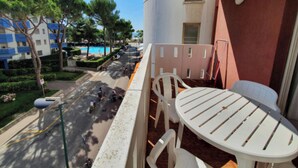 Un balcón con una mesa redonda y sillas, con vista a una calle con coches aparcados y una piscina al fondo.